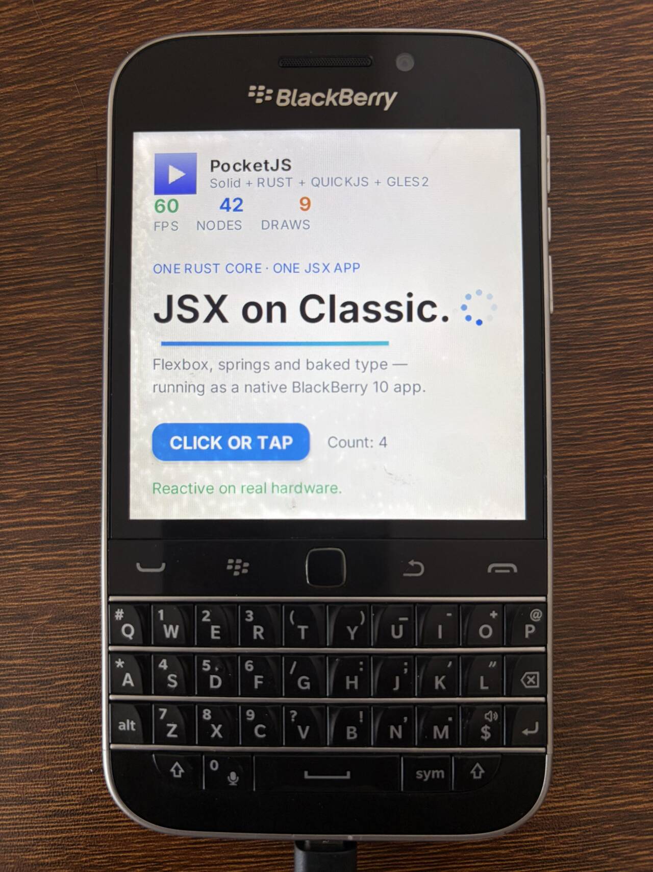 A real BlackBerry Classic on a wooden desk, its square screen running the PocketJS Hero demo: a PocketJS header with 60 FPS / 42 NODES / 9 DRAWS counters and 'ONE RUST CORE · ONE JSX APP', the headline 'JSX on Classic.', body text 'Flexbox, springs and baked type — running as a native BlackBerry 10 app.', a blue 'CLICK OR TAP' button, 'Count: 4', and 'Reactive on real hardware.' Below the square screen are the physical QWERTY keyboard and the tool belt with its optical trackpad.